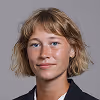 Portrait of a young woman with short blonde hair wearing a dark blazer and white shirt, smiling slightly against a gray background.